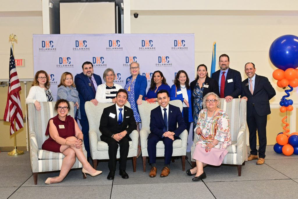 A formal group photo of Delaware Hispanic Commission members standing and sitting in front of a branded backdrop.