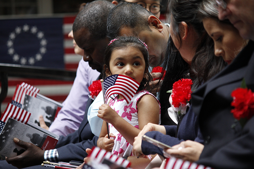 A young girl holding a small U.S. flag looks ahead during a naturalization ceremony, surrounded by new American citizens holding flags and red carnations.